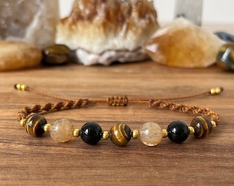 May include: A brown cord bracelet with gold beads and gemstone beads. The beads are tiger's eye, clear quartz, and black onyx.