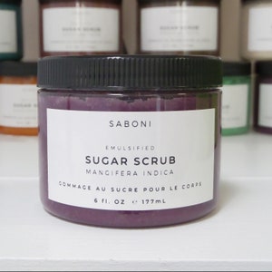 May include: A purple sugar scrub jar with a black lid. The label reads "SABONI EMULSIFIED SUGAR SCRUB MANGIFERA INDICA GOMMAGE AU SUCRE POUR LE CORPS 6 fl. oz e 177mL".
