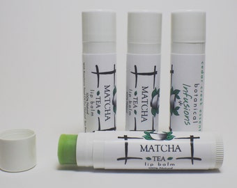 Può includere: Tre tubetti bianchi di balsamo per labbra al tè matcha, 100% naturale, con accenti di foglie verdi e il testo "Matcha Tea Lip Balm 100% Natural" sulle etichette. Un tubetto è aperto, rivelando un bastoncino di balsamo per labbra verde.