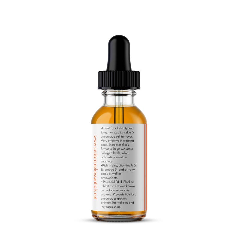 Op de afbeelding: Een doorzichtige glazen fles met een zwarte druppelaar bovenop, gevuld met een licht amberkleurige vloeistof. De fles is gelabeld met de tekst "Great for all skin types. Enzymes exfoliate skin & encourage cell turnover. Very effective in treating acne. Increases skin's firmness, helps maintain collagen levels, which prevents premature sagging. Rich in zinc, vitamins A & E, omega 3- and 6- fatty acids as well as antioxidants. Powerful DHT Blockers inhibit the enzyme known as 5-alpha reductase enzyme. Prevents hair loss, encourages growth, protects hair follicles and increases shine." en de website "www.cedarcreekessentials.net".