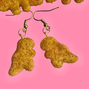 May include: Gold-colored dinosaur-shaped earrings with gold-tone ear wires. The earrings are set against a bright pink background, with additional dinosaur shapes scattered around the frame. The earrings are a fun and quirky accessory.