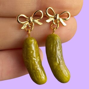 May include: A pair of dangle earrings featuring gold-toned metal bows at the top, each suspending a realistic, textured green pickle charm. The pickles have a bumpy, cylindrical shape with a glossy finish, hanging from small gold jump rings.