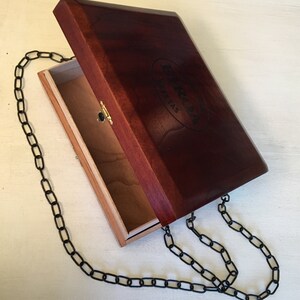 Könnte beinhalten: Eine rechteckige Holzkiste mit einem Klappdeckel, der tiefrotbraun gebeizt ist. Der Deckel ist leicht geöffnet und gibt einen hellfarbigen Innenraum frei. Eine schwarze Kette ist an der Box befestigt, was darauf hindeutet, dass sie als Handtasche getragen werden kann. Die Box hat einen goldenen Verschluss.