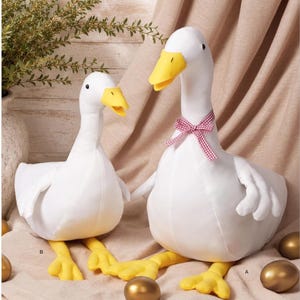 May include: A Simplicity S3296 sewing pattern envelope featuring two white fabric goose plushies with yellow beaks and feet. The larger goose wears a red and white gingham ribbon around its neck. The design is labeled easy-to-sew and designed in USA.