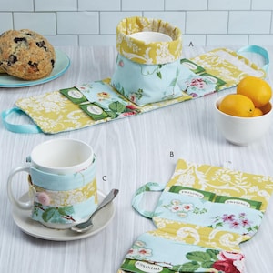 May include: A Kwik Sew K4313 sewing pattern for tea time accessories. The image displays fabric tea bag holders and mug cozies in light blue and yellow floral patterns, featuring pockets for tea bags and a pink button closure on the mug cozy.