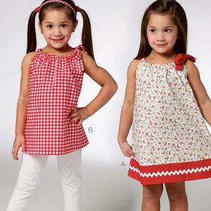 May include: Kwik Sew pattern K3934 for toddlers' clothing. The image shows two young girls wearing handmade dresses and tops. One girl wears a red and white gingham top with white pants. The other wears a floral dress.