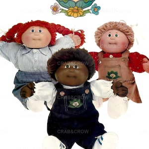 May include: Three Cabbage Patch Kids dolls with different skin tones and hairstyles. One doll wears a blue and white striped shirt and denim overalls. Another doll wears a red shirt and brown overalls. The third doll wears a dark blue overall. The Cabbage Patch Kids logo is at the top.