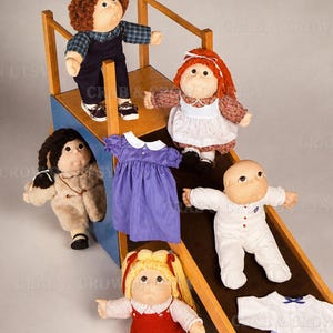 May include: Five soft-sculptured dolls with yarn hair and fabric clothing arranged on a wooden play structure. The dolls wear various outfits including overalls, dresses, and pajamas. A purple dress and a white and blue outfit are displayed separately.