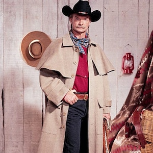 May include: A beige full-length duster coat with a shoulder cape, worn over a red button-down shirt and dark blue jeans. The outfit includes a black cowboy hat, a patterned neckerchief, a brown leather belt, and black leather boots.