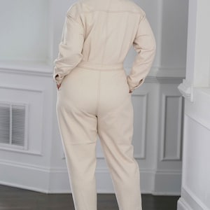 Peut inclure: Une combinaison cr&egrave;me &agrave; manches longues et une ceinture. La combinaison a un col et une fermeture boutonn&eacute;e. Le mannequin porte des talons hauts marrons.