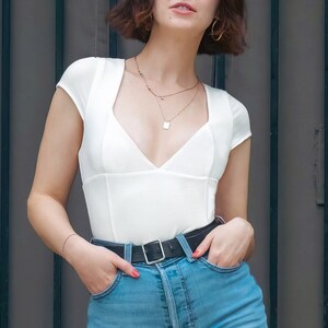 May include: A woman wearing a white, fitted, v-neck top with short sleeves and a black belt. She is also wearing light blue denim shorts.