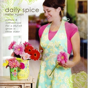 May include: A teal blue halter apron with a floral pattern. The apron has a V-neck and a tie at the back. The model is wearing a pink shirt and holding a bouquet of flowers.