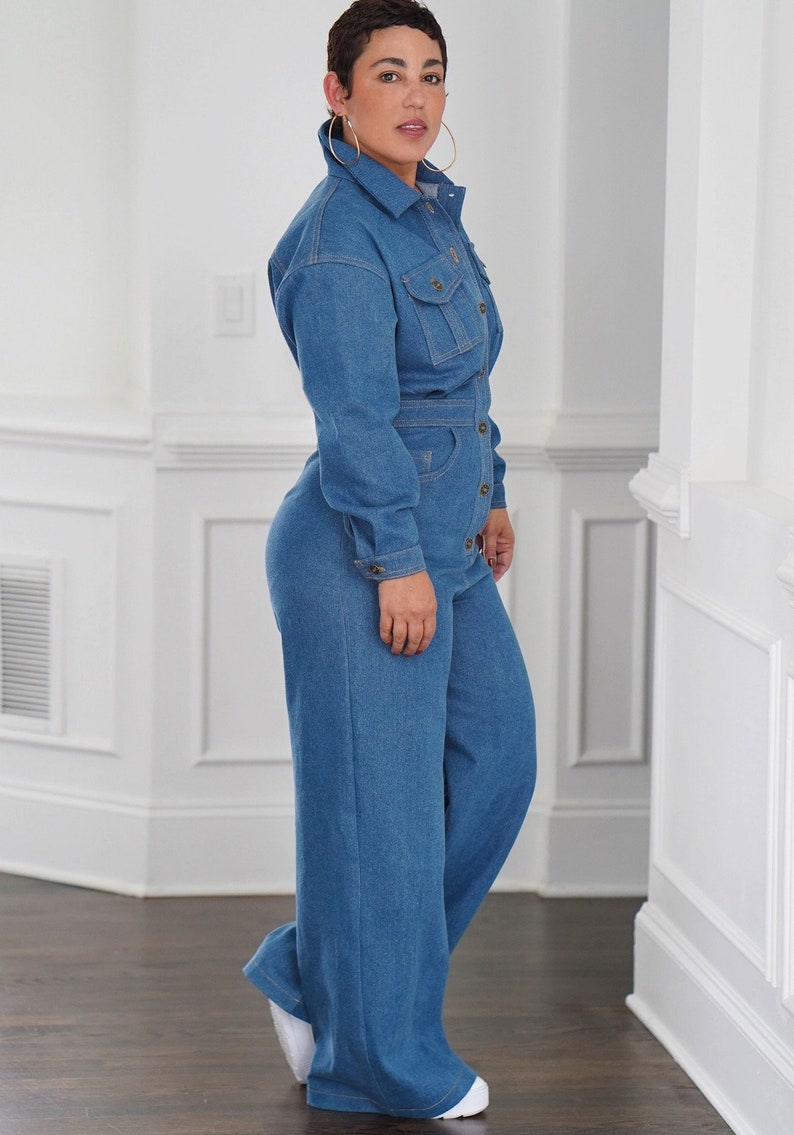 Peut inclure: Une femme portant une combinaison en jean bleu &agrave; manches longues et &agrave; fermeture boutonn&eacute;e. La combinaison a une coupe ample et une ceinture &agrave; la taille.