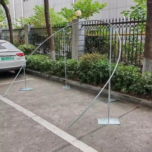 May include: A silver metal archway structure, composed of curved and straight metal rods, is set up outdoors. The arch is supported by rectangular base plates. The background includes a car, greenery, and a fence.