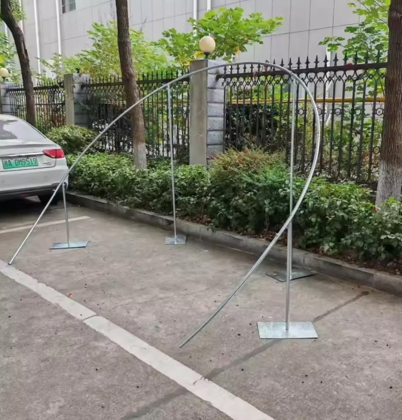 May include: A silver metal archway structure, composed of curved and straight metal rods, is set up outdoors. The arch is supported by rectangular base plates. The background includes a car, greenery, and a fence.