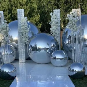 May include: A wedding ceremony backdrop with silver mirrored spheres of various sizes. The spheres are arranged in a semi-circle with white floral arrangements on either side.