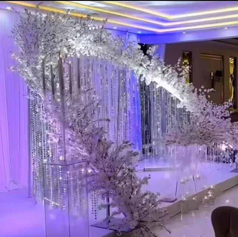 May include: A decorative wedding arch with white floral arrangements and hanging crystal strands. The arch is set against a backdrop of shimmering curtains and is illuminated by soft lighting, creating an elegant ambiance.