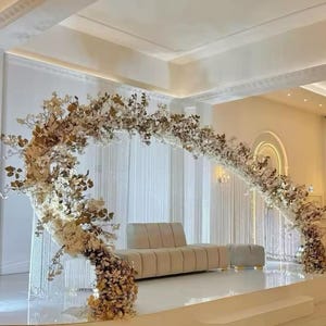 May include: A wedding stage setup featuring a beige sofa and ottoman set. A large floral arch with white and gold flowers frames the seating area. The stage is white, with a backdrop of white curtains.