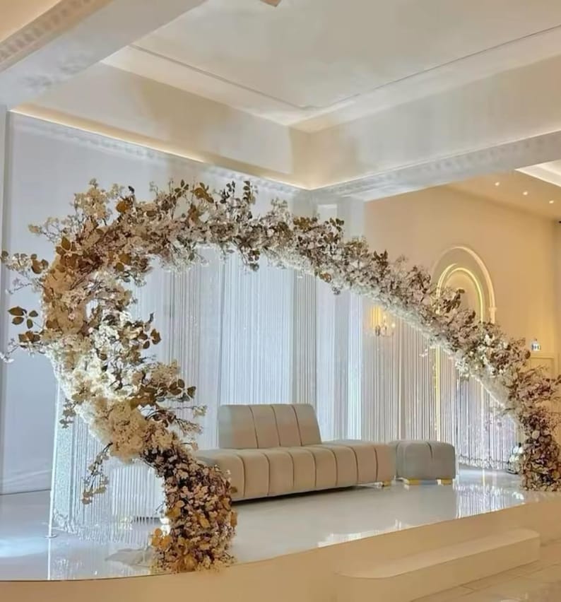 May include: A wedding stage setup featuring a beige sofa and ottoman set. A large floral arch with white and gold flowers frames the seating area. The stage is white, with a backdrop of white curtains.