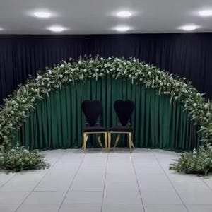 May include: A wedding ceremony setup featuring two black chairs with heart-shaped backs and gold legs. A lush floral arch of white flowers and greenery frames a dark green curtain backdrop. The scene is illuminated by overhead lights.