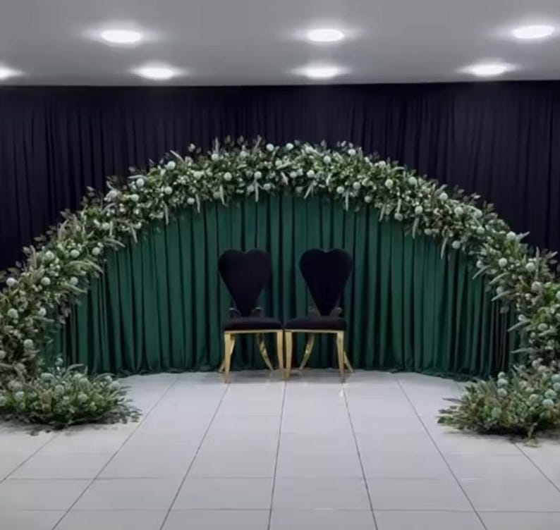 May include: A wedding ceremony setup featuring two black chairs with heart-shaped backs and gold legs. A lush floral arch of white flowers and greenery frames a dark green curtain backdrop. The scene is illuminated by overhead lights.