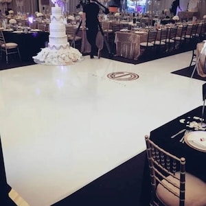 May include: A white dance floor with a black border and a circular design in the center. The floor is in a large room with tables and chairs set up around it. There is a white wedding cake on a table in the background.