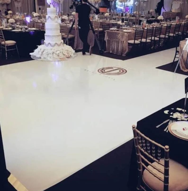 May include: A white dance floor with a black border and a circular design in the center. The floor is in a large room with tables and chairs set up around it. There is a white wedding cake on a table in the background.