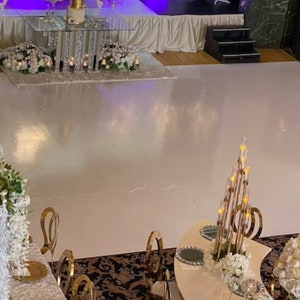 May include: A white dance floor with gold chairs and white floral arrangements. The dance floor is surrounded by a white tablecloth and a gold table with a white cake on top.