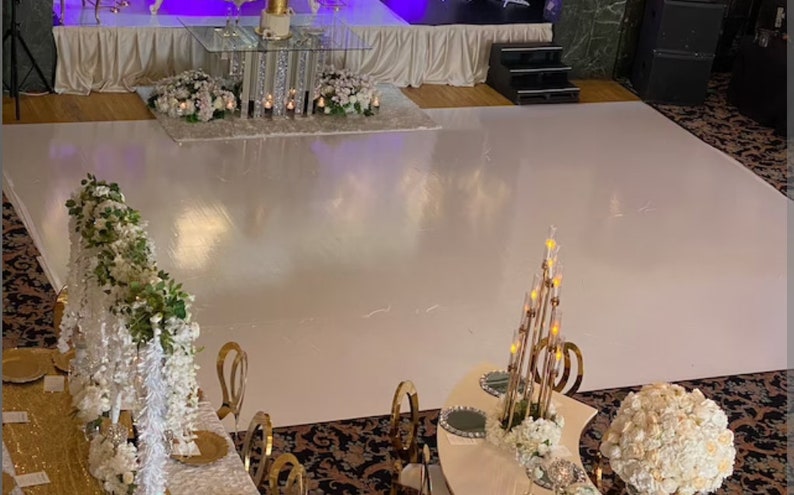 May include: A white dance floor with gold chairs and white floral arrangements. The dance floor is surrounded by a white tablecloth and a gold table with a white cake on top.