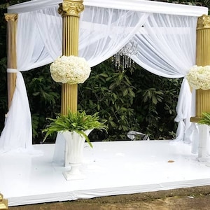 May include: White wedding ceremony archway with gold columns and white fabric draped over the top. The archway is decorated with white flowers and greenery.