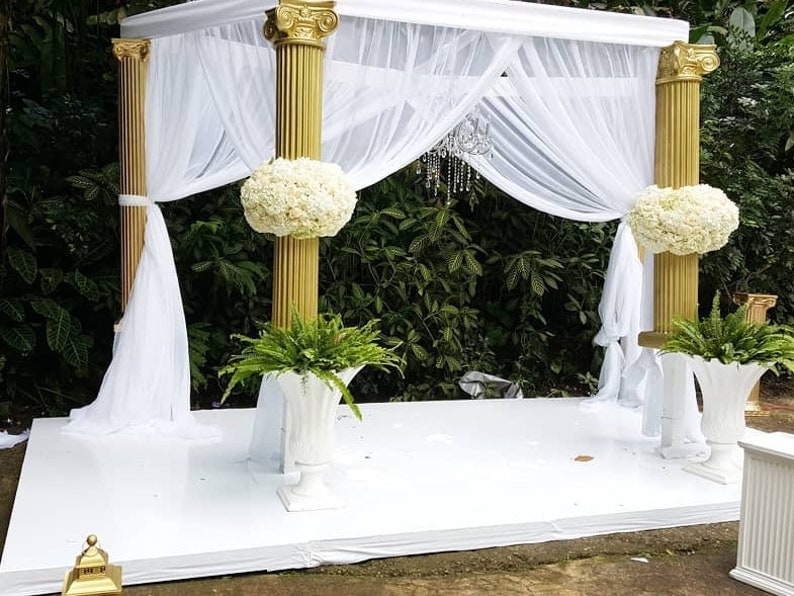 May include: White wedding ceremony archway with gold columns and white fabric draped over the top. The archway is decorated with white flowers and greenery.