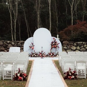 May include: Outdoor wedding ceremony setup with white chairs lining the aisle leading to a white backdrop with a large, round, white arch and floral arrangements in shades of pink and burgundy.