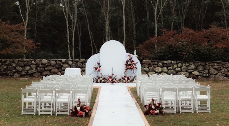 May include: Outdoor wedding ceremony setup with white chairs lining the aisle leading to a white backdrop with a large, round, white arch and floral arrangements in shades of pink and burgundy.