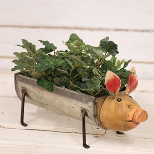 May include: A decorative planter shaped like a pig, made of metal and wood. The planter has a trough for plants, filled with green foliage. The pig has a painted face with red-tipped ears and a pink snout. The planter is on four metal legs.