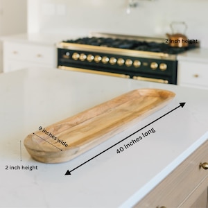 May include: A light-colored wooden serving tray, 40 inches long, 9 inches wide, and 2 inches high. The tray has a natural wood grain and a curved, boat-like shape. It is displayed on a white countertop.