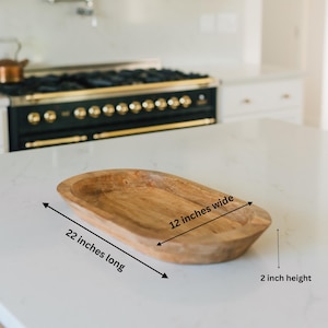 May include: A wooden, oval-shaped bowl, 22 inches long, 12 inches wide, and 2 inches high. The bowl has a natural wood grain finish. The kitchenware is displayed on a white countertop.