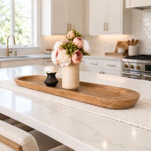 May include: A long, oval-shaped wooden dough bowl sits on a white surface. Inside the bowl, there is a beige cylindrical vase filled with a bouquet of pink and white artificial peonies and a small, dark, round vase holding a white flower.