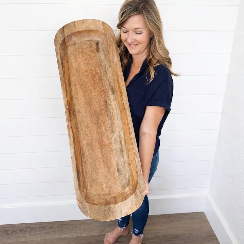 May include: A large, rectangular wooden dough bowl with rounded ends. The bowl is made of light brown wood with visible grain and a carved interior. The bowl is being held by a person.