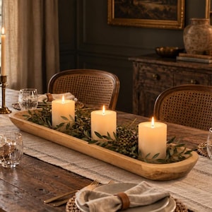 May include: A long, shallow, natural wood dough bowl centerpiece containing three lit white pillar candles nestled among green foliage and small berries, arranged on a wooden dining table with a linen runner and place settings.