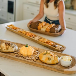 May include: Wooden serving trays filled with bread, crackers, and a decorative pumpkin. One tray holds various types of bread, while another features crackers and a small pumpkin. A small bowl of butter is also present.
