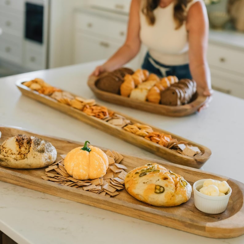 May include: Wooden serving trays filled with bread, crackers, and a decorative pumpkin. One tray holds various types of bread, while another features crackers and a small pumpkin. A small bowl of butter is also present.