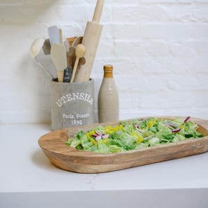 May include: A wooden serving bowl filled with a fresh salad of green lettuce, red onion, and yellow peppers. A ceramic utensil holder with the text "UTENSILS Paris, France 1893" and a bottle of dressing are in the background.