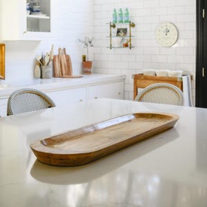May include: A long, oval-shaped wooden serving tray with a light brown finish. The tray has a shallow, recessed center and rounded edges. It is placed on a white countertop in a kitchen setting.