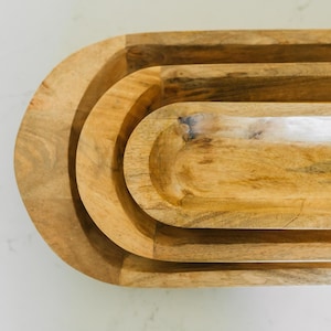 May include: A set of three wooden oval-shaped trays nested together. The trays are made of light-colored wood with visible grain and a smooth, polished finish. The trays are designed to nest inside each other.