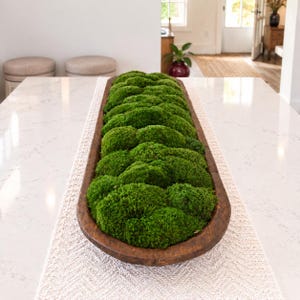 May include: A long, narrow, dark brown wooden dough bowl filled with vibrant green preserved moss. The bowl has a smooth, curved edge and a rustic texture, containing multiple rounded clusters of dense, textured moss that fill the entire interior.