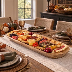 May include: A long, oval-shaped wooden dough bowl filled with an assortment of crackers, sliced meats, cheeses, grapes, and small glass containers of dips, arranged as a charcuterie board on a dining table with place settings.