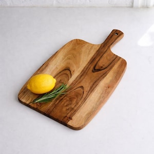 May include: A wooden cutting board with a handle, featuring a bright yellow lemon and a sprig of rosemary. The board has a natural wood grain pattern and is set against a white background. This kitchen item is ideal for food preparation.