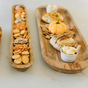 May include: Two wooden serving trays with food. One tray holds an assortment of crackers. The other tray has bread, a pumpkin-shaped butter, and a small bowl of butter. The trays are on a white surface.