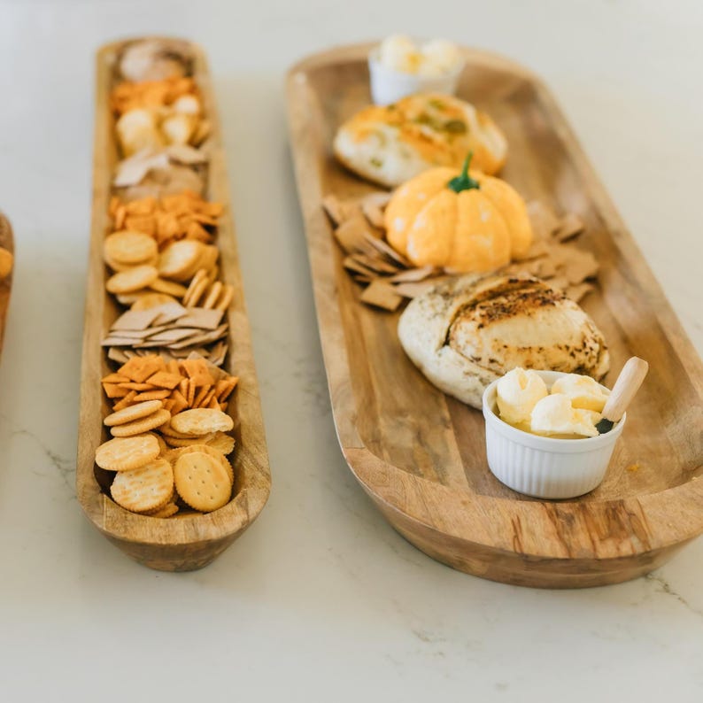 May include: Two wooden serving trays with food. One tray holds an assortment of crackers. The other tray has bread, a pumpkin-shaped butter, and a small bowl of butter. The trays are on a white surface.