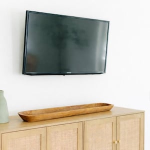 May include: A black flat-screen television mounted on a white wall above a wooden cabinet. A long, wooden, oval-shaped bowl sits on top of the cabinet. The cabinet has woven panels on the doors.